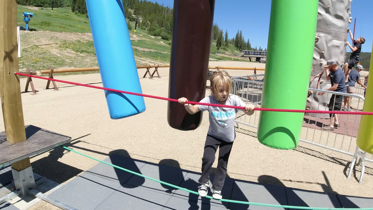 The child moves farther along the Winter Park obstacle course, gripping the red rope as blue, brown, green, and yellow foam cylinders hang nearby.