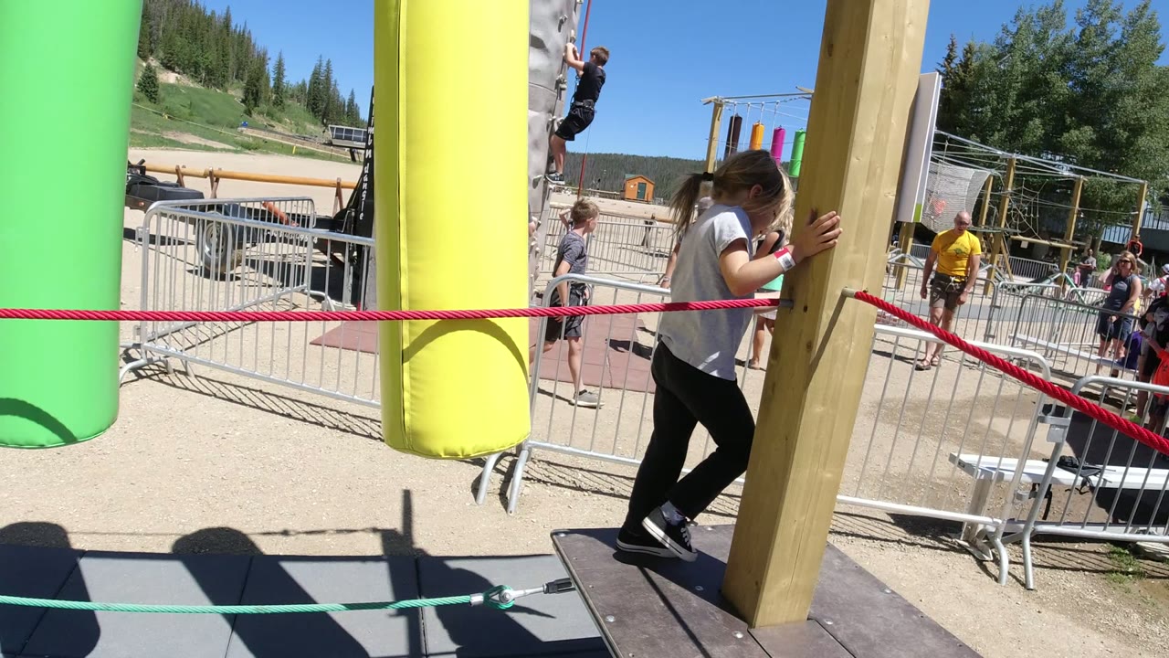 A child steps along the rope obstacle in Winter Park, Colorado, partly blocked by a wood support post, with colorful hanging elements and the climbing wall behind.