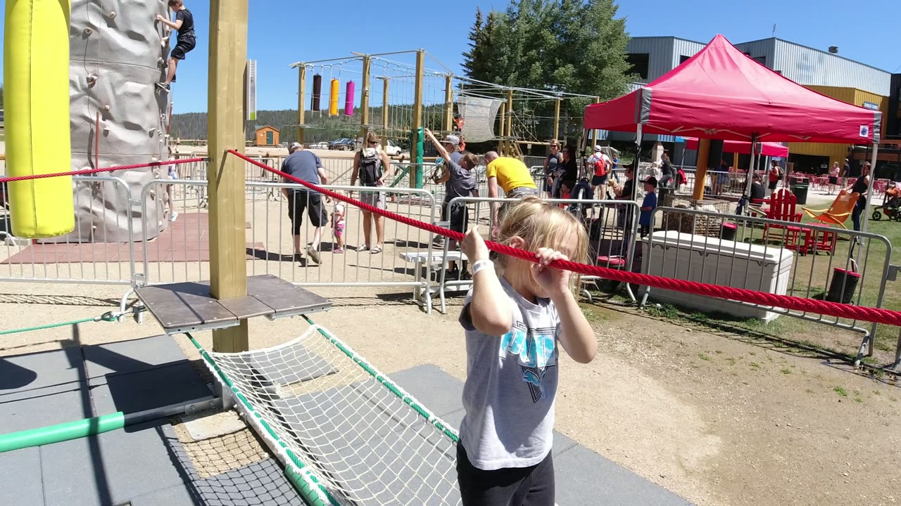 The child moves closer to the camera while gripping the red rope, with the net bridge, yellow post, red canopy, barriers, and background visitors visible in Winter Park, Colorado.