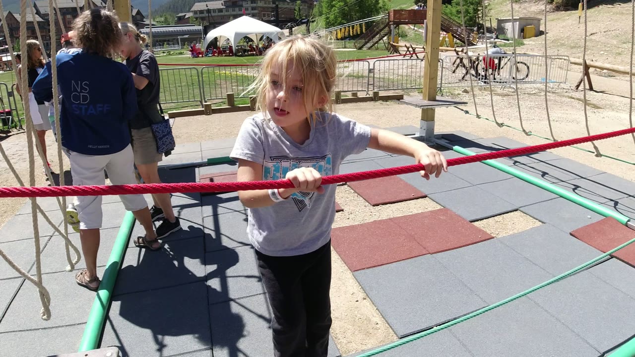 The child faces toward the camera in Winter Park, Colorado while holding the red rope, with adults, a white tent, bicycles, and hillside buildings in the background.