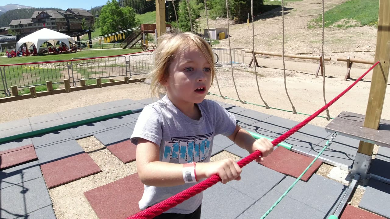 The girl faces forward while holding the red rope with both hands, with a white tent, rails, bare slope, trees, and distant buildings visible in Winter Park, Colorado.