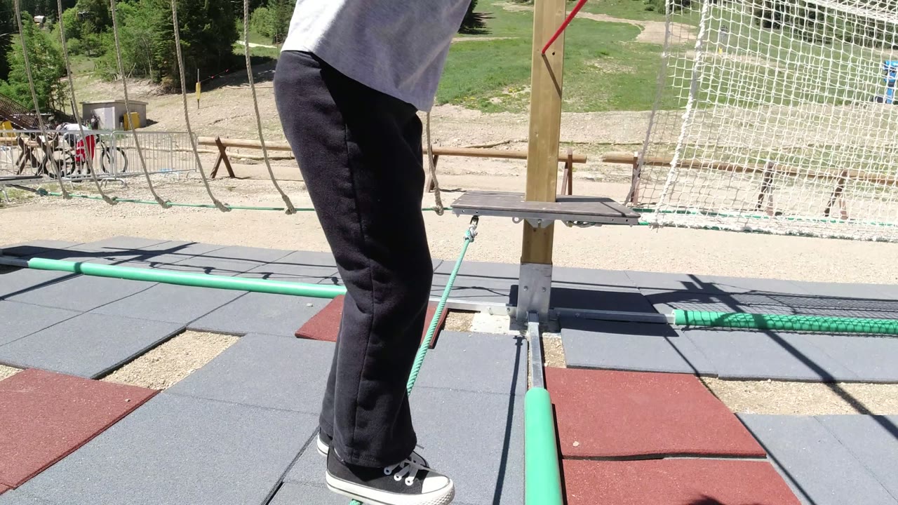 Opening frame in Winter Park, Colorado showing a child partly cropped at the left edge on a ropes playground, with padded ground tiles, a red rope, a green balance rope, netting, and sunny mountain recreation grounds behind.