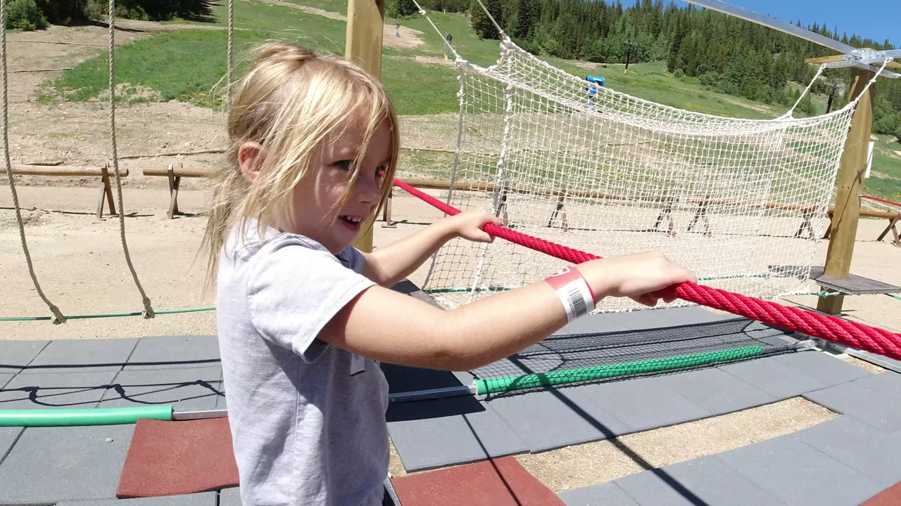 A blond child in Winter Park, Colorado faces the camera while gripping a red rope on a playground course, with a rope net, wooden supports, padded flooring, open ground, trees, and bright blue sky visible.