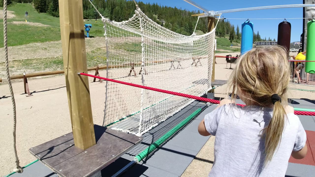 The child remains centered in the frame in Winter Park, Colorado, with both hands near the red rope and the white net bridge stretched ahead in bright sun.