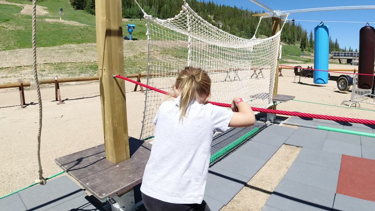 A child in Winter Park, Colorado holds a red rope from behind beside white netting, a wooden post, padded flooring, sandy ground, and green slopes.