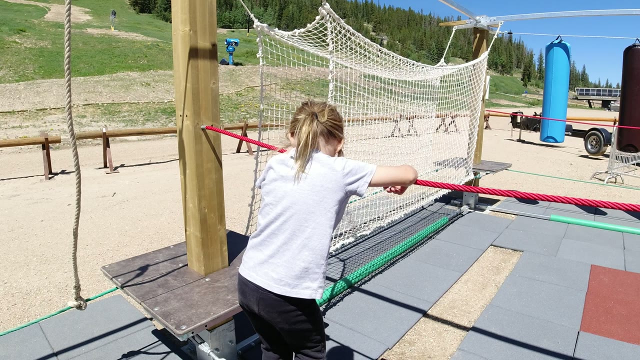 The child leans farther along the red rope in Winter Park, Colorado, with the white net obstacle, wooden support, and mountain slope behind.