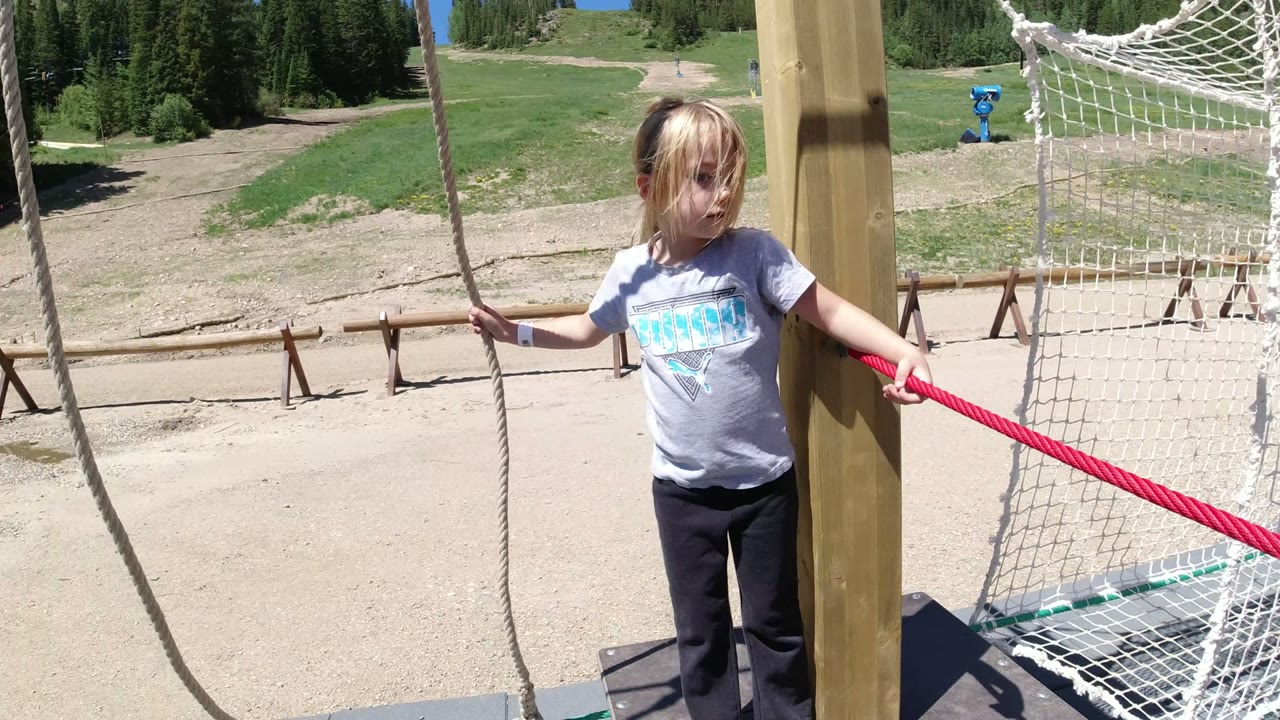 Child holding ropes beside a netted play bridge on a sunny mountain slope.