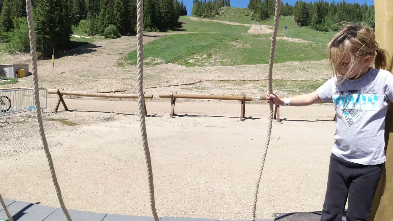 In Winter Park, Colorado, the child stands between hanging ropes, looking downward while the open tan ground, green slope, and dark evergreen treeline fill the background.