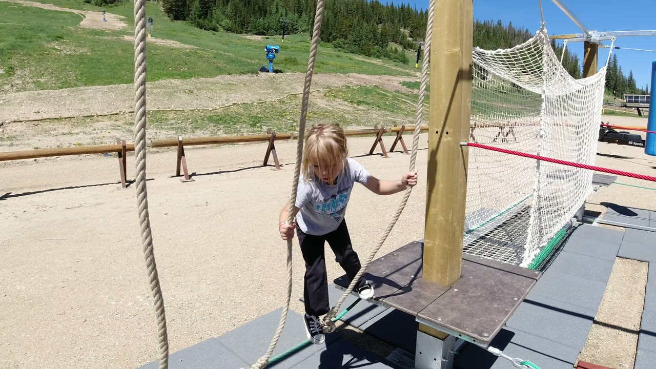 The child shifts footing on the playground platform while gripping the ropes, with gravel paths, hillside grass, and pine trees behind.