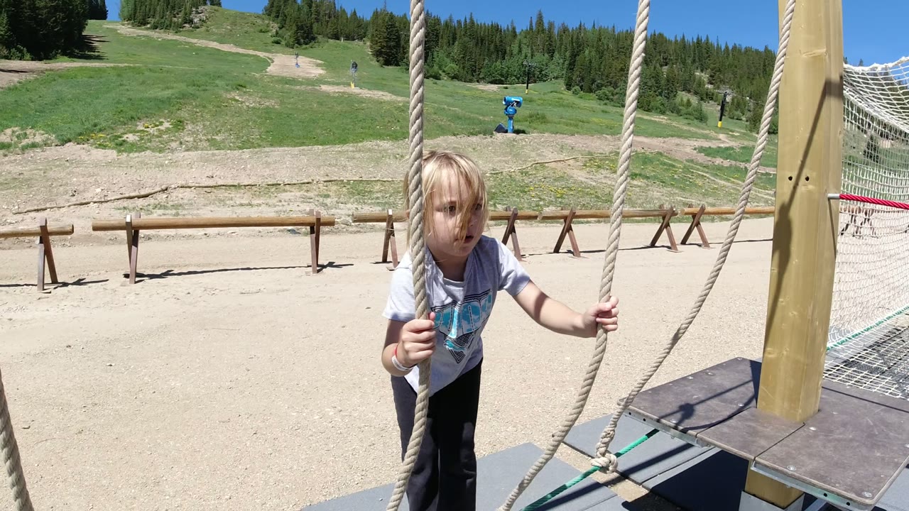 The girl faces closer toward the camera while holding both rope lines, with the rope platform and hillside play area still in view in Winter Park, Colorado.