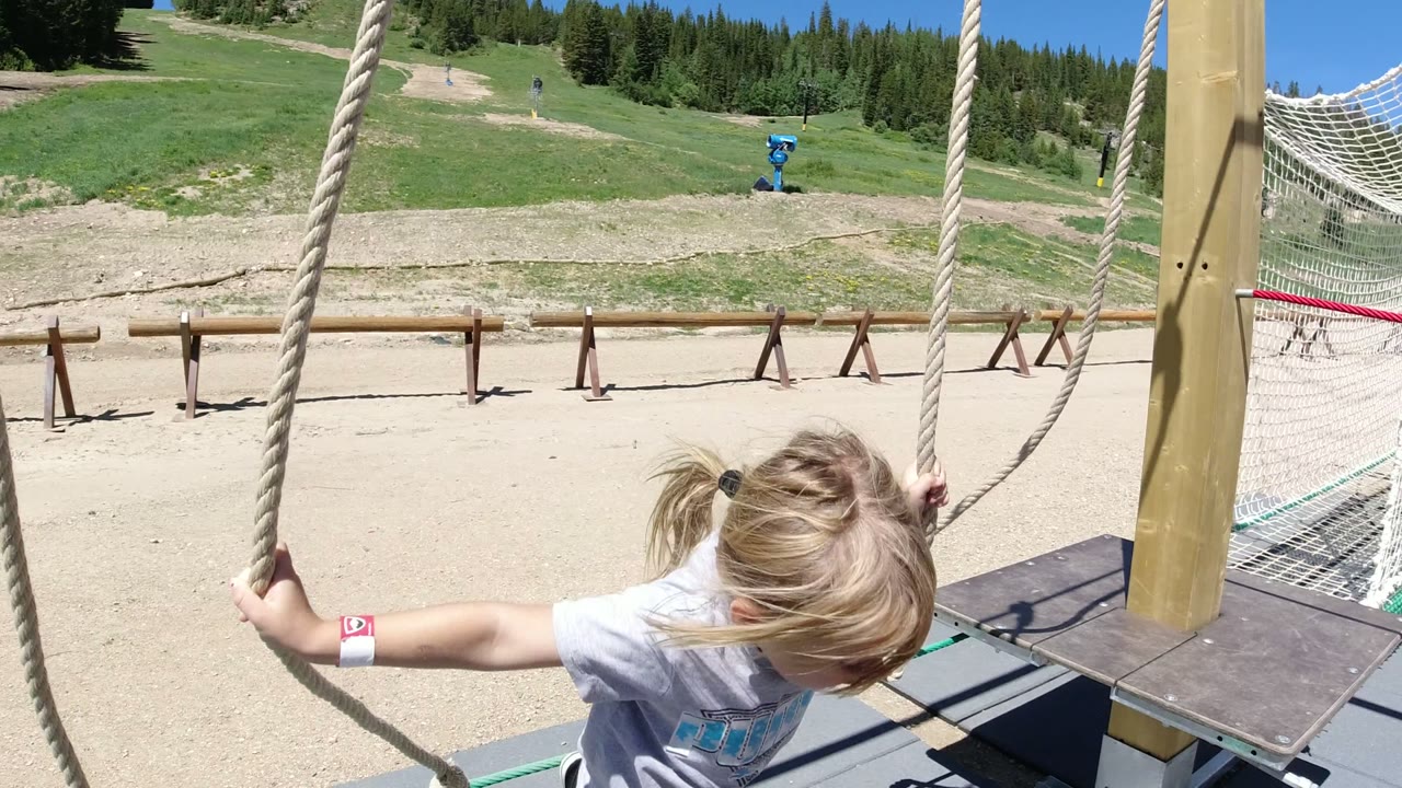 At 0:00 in Winter Park, Colorado, a blond child holds a thick rope near a small wooden platform, with dirt paths, grass, and evergreen trees in the background.