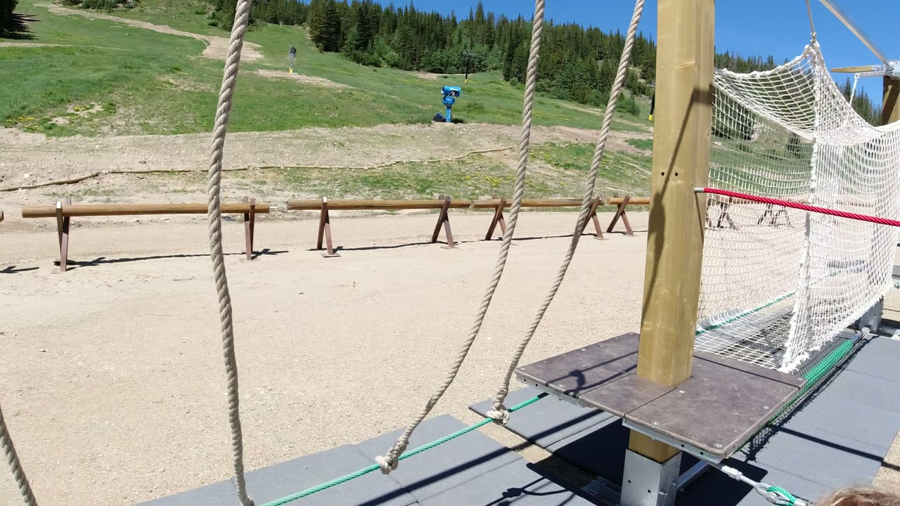 At 0:01 in Winter Park, Colorado, the rope course is shown without visible people, with hanging ropes, a wooden platform, white netting, dirt paths, grass, and trees beyond.