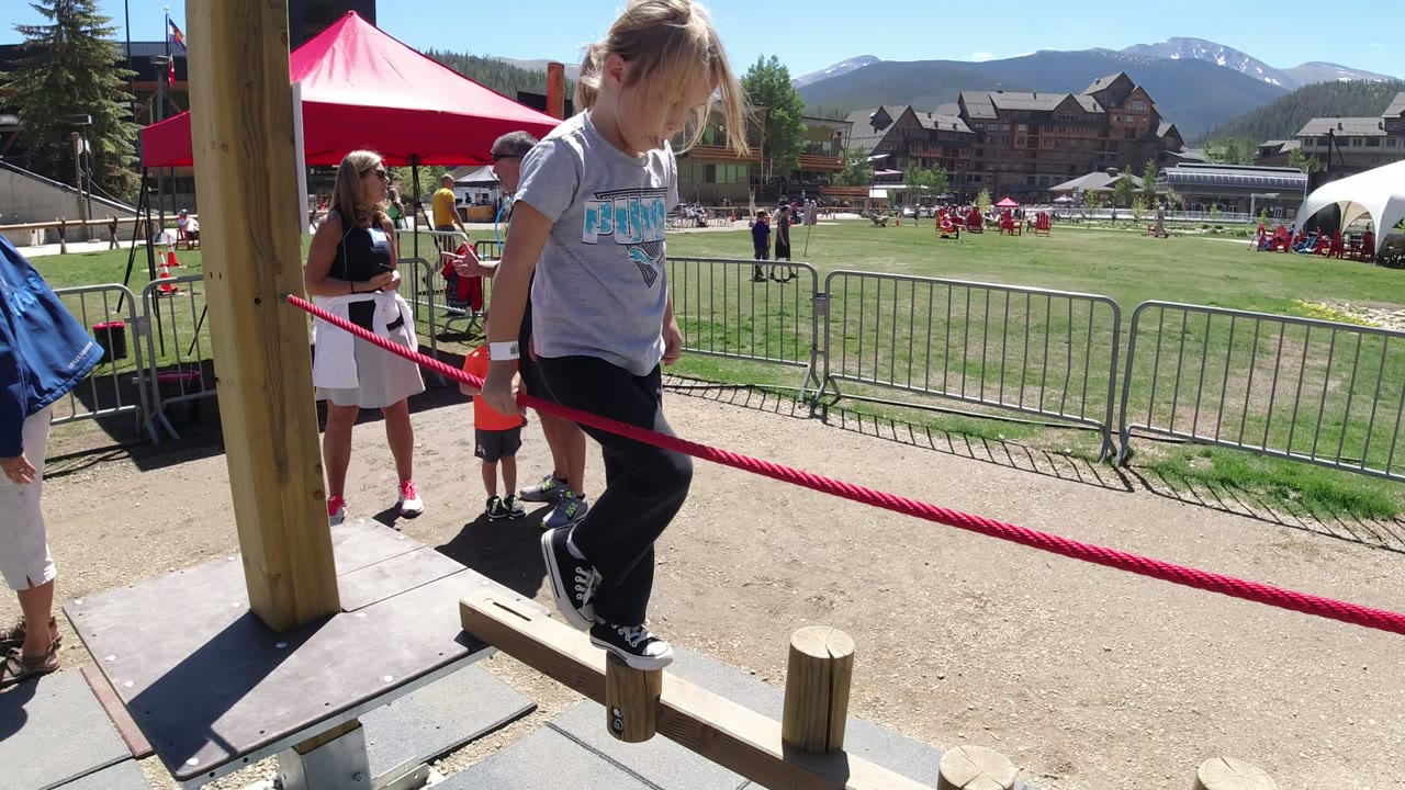 At the start of the Winter Park, Colorado clip, a child stands on a wooden balance post with both hands near a red rope as adults and event fencing sit behind her.