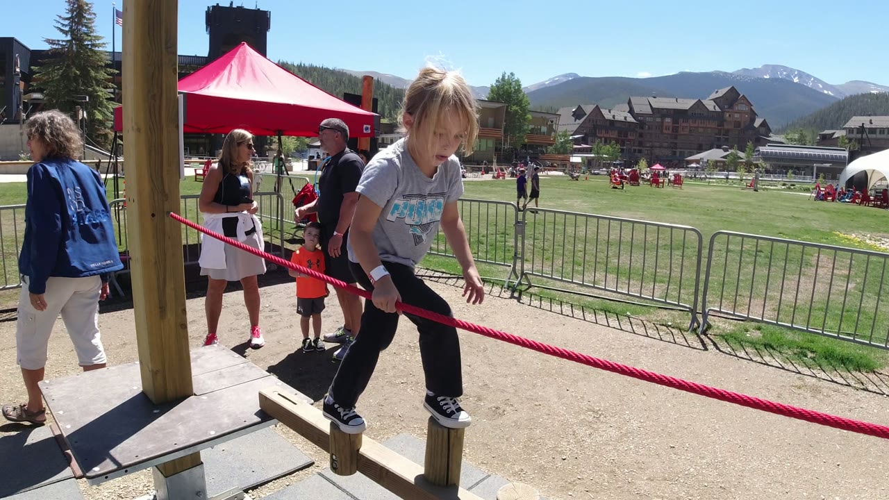 A child in Winter Park, Colorado leans forward on the low wooden posts, using the red rope for support while spectators stand near a red tent and metal barriers.