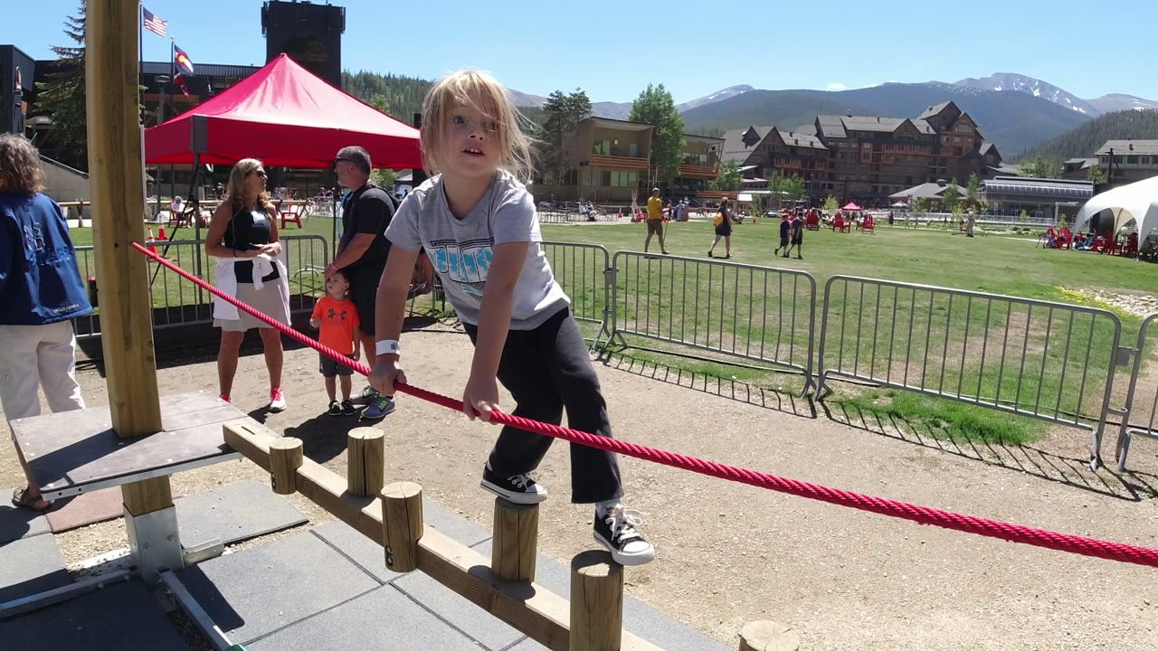 Child holding a red rope on a wooden balance obstacle at an outdoor event.