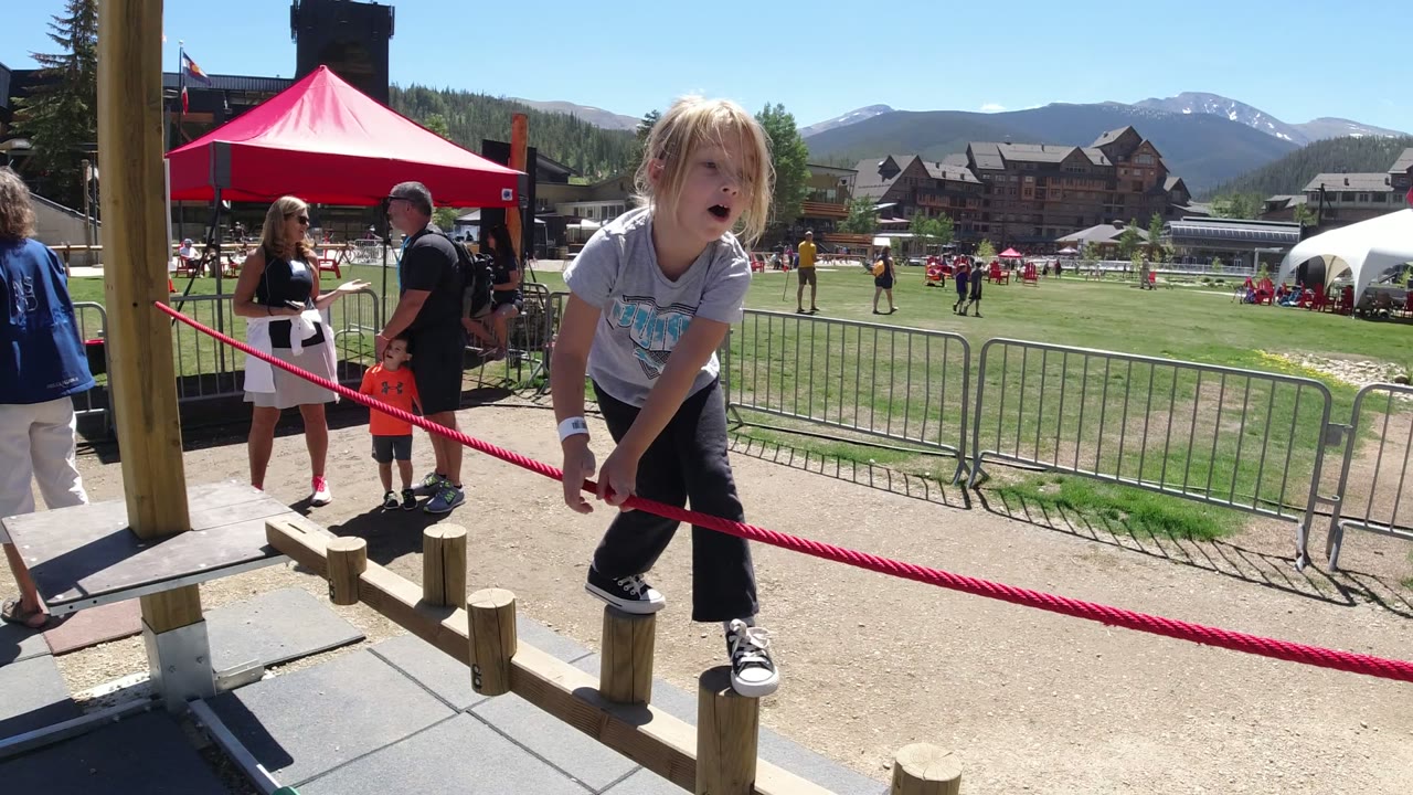 The child leans forward on the wooden posts in Winter Park, Colorado, with a red tent, several adults, a small child, barricades, lawn, buildings, and distant mountains in frame.