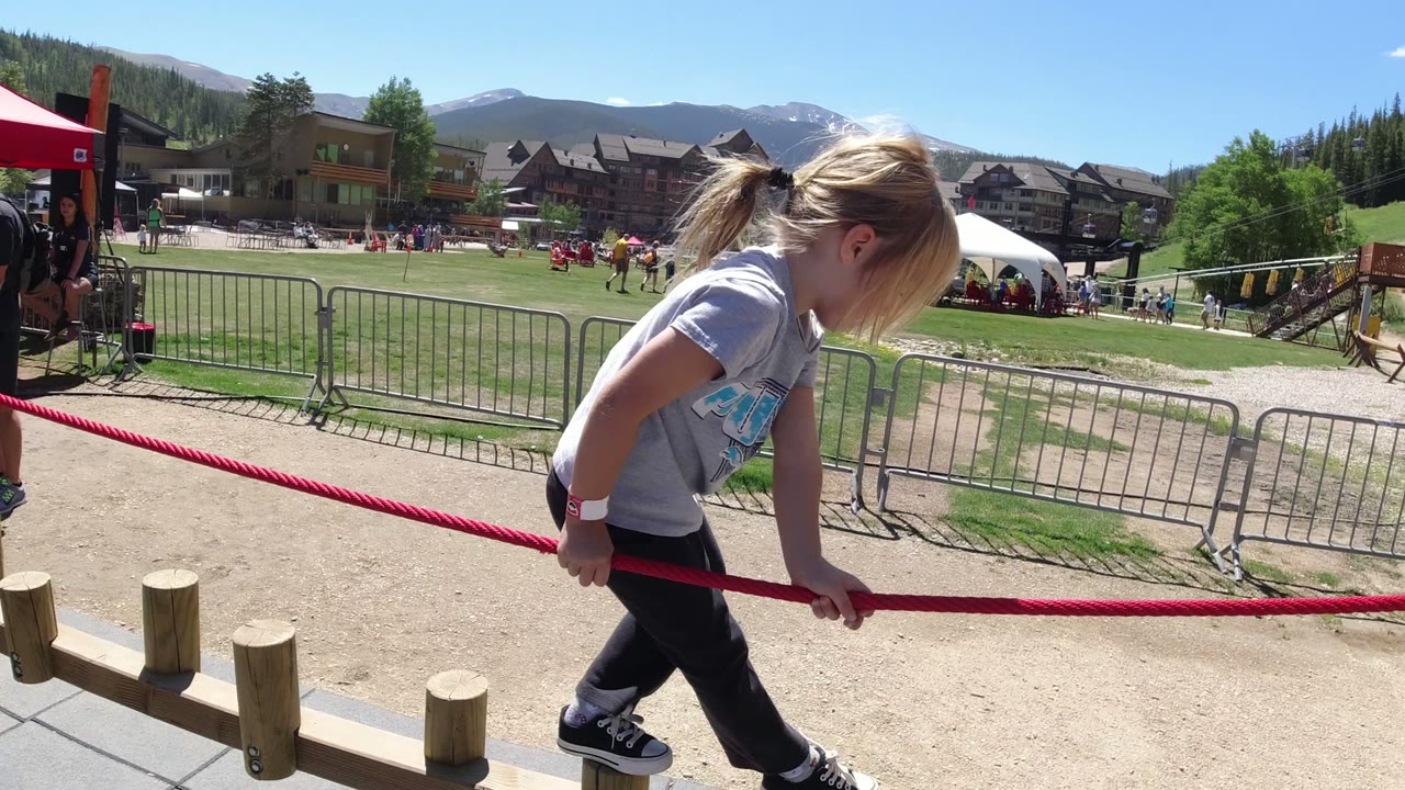 The girl leans forward with both hands on the red rope in Winter Park, Colorado, with a fenced grass area, tents, and mountain buildings in the background.