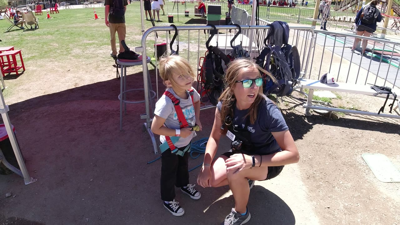 Woman kneeling beside a young boy in a harness at an outdoor recreation area.