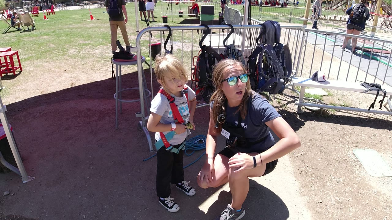 A kneeling woman looks upward beside the boy in a harness, with shaded dirt in the foreground and metal fencing, bags, and grass behind them in Winter Park, Colorado.