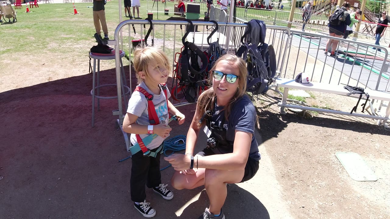 Child in a red harness stands beside a kneeling woman near outdoor gear.
