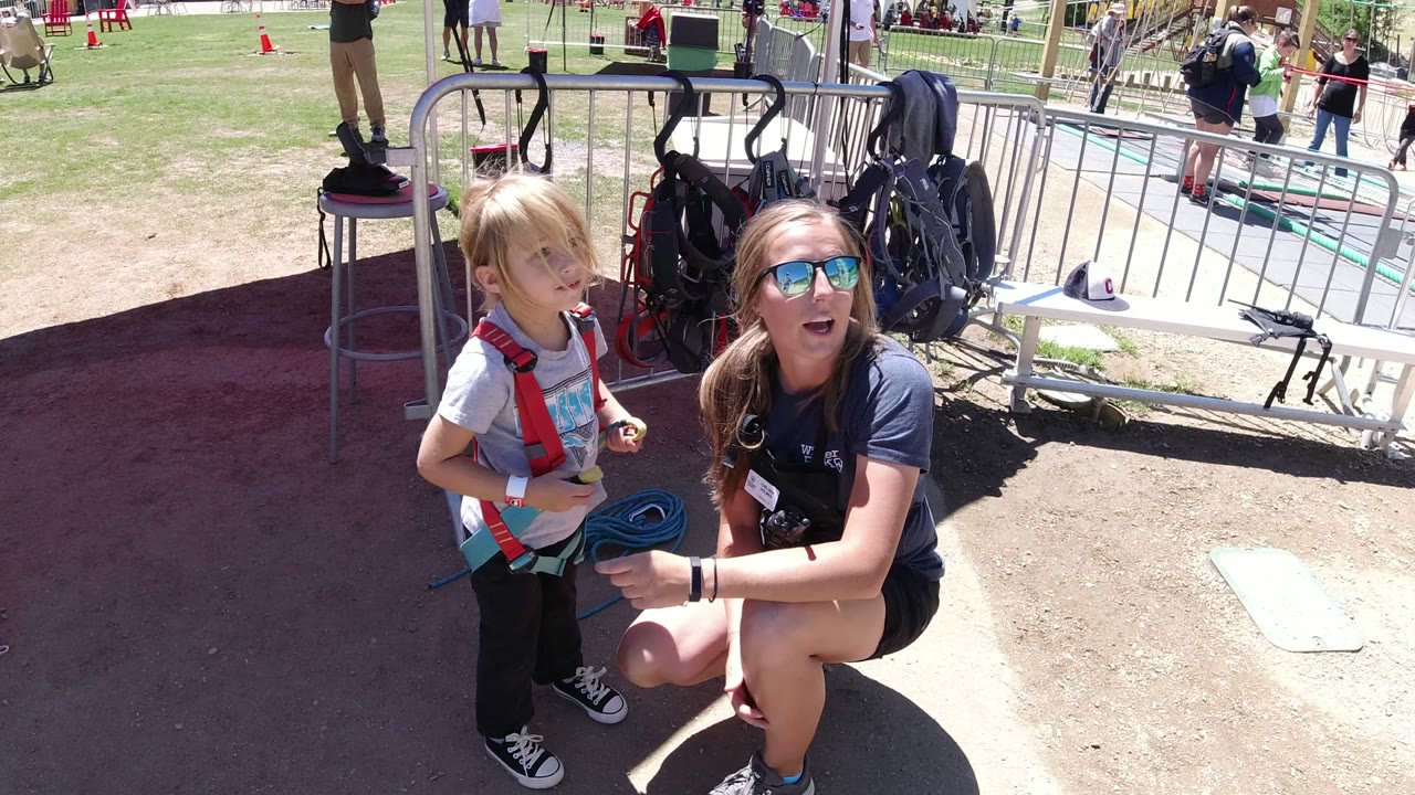 In Winter Park, Colorado, the kneeling woman looks upward while the child in a red harness stands next to her near coiled rope and activity barriers.