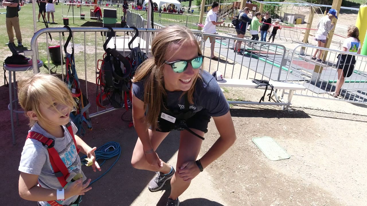 Female helper kneels beside a young girl in a harness at an outdoor activity area.