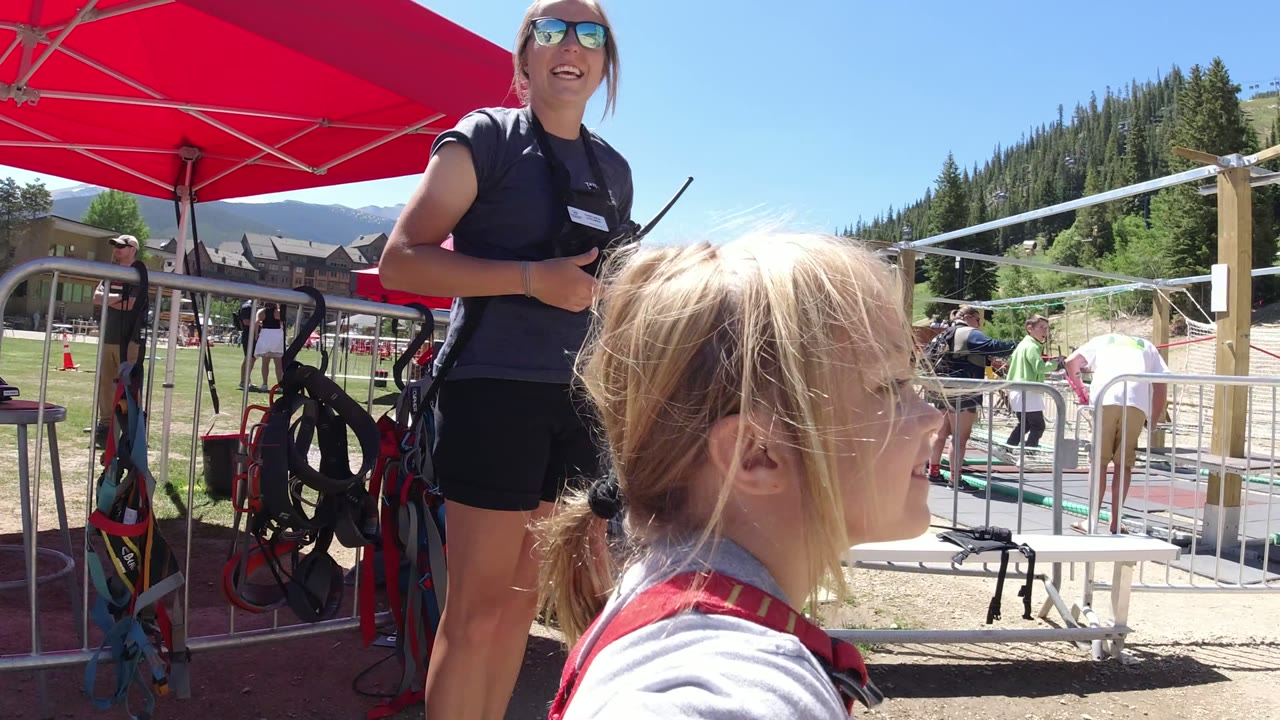 A young girl in red overalls looks toward a black-clad attendant under a red canopy at an outdoor park in Winter Park, Colorado.