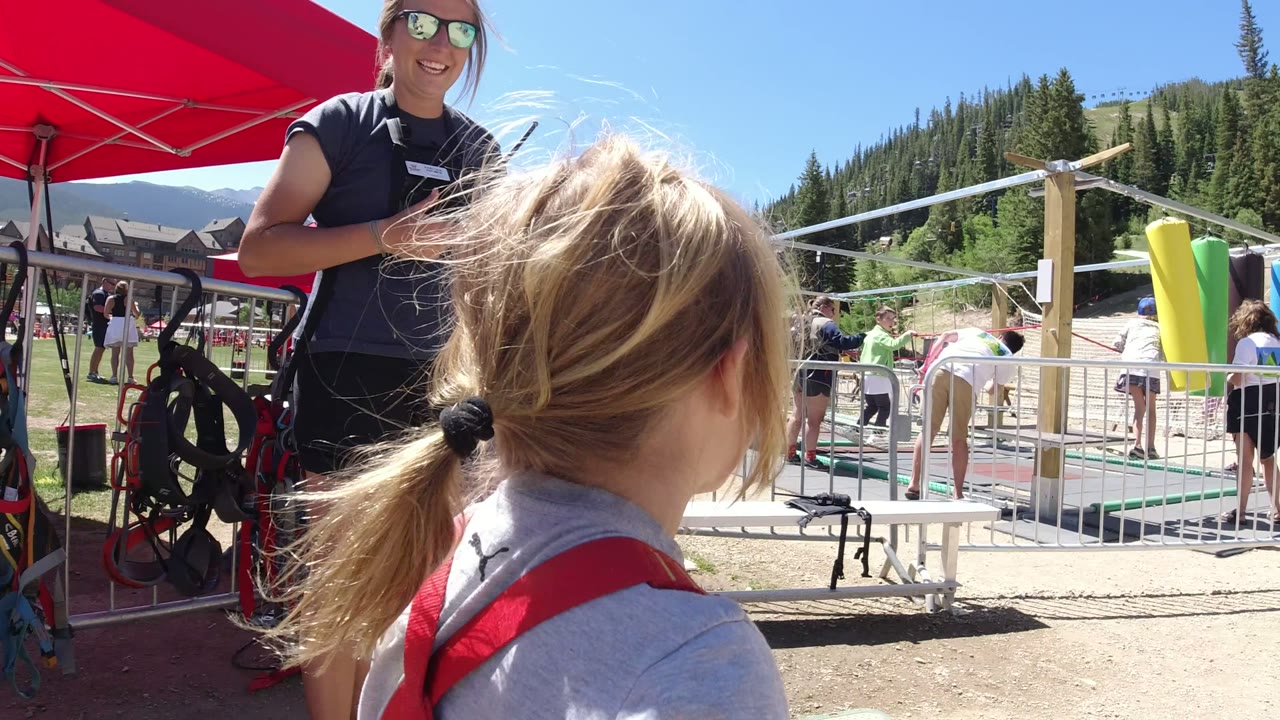 The girl is seen from behind with her ponytail and red overall straps visible as the attendant stands by equipment and railings in Winter Park, Colorado.