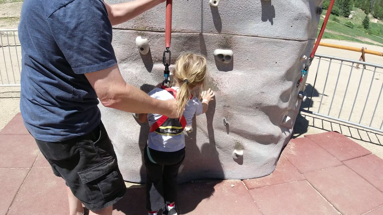 In Winter Park, Colorado, an adult in a blue shirt stands behind a blonde child in a harness at the lower holds of an outdoor climbing wall.