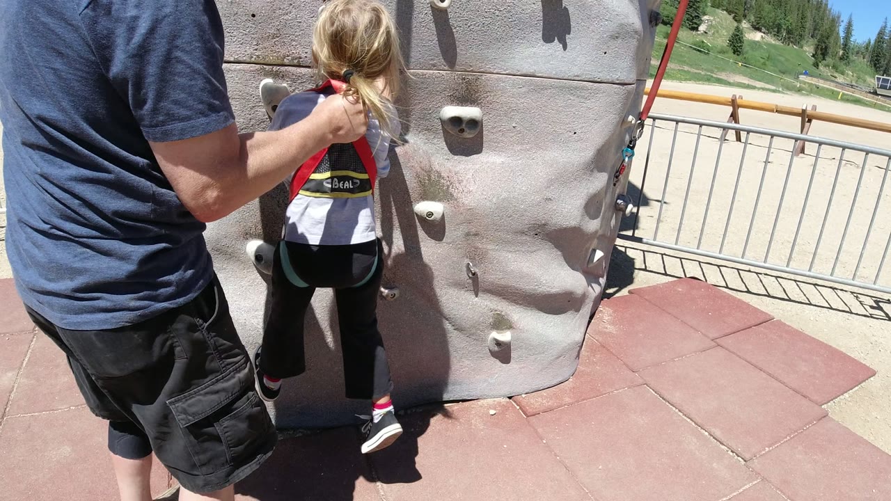 In Winter Park, Colorado, the child has one foot on a low climbing hold while the adult keeps a hand near the harness and shoulder area.