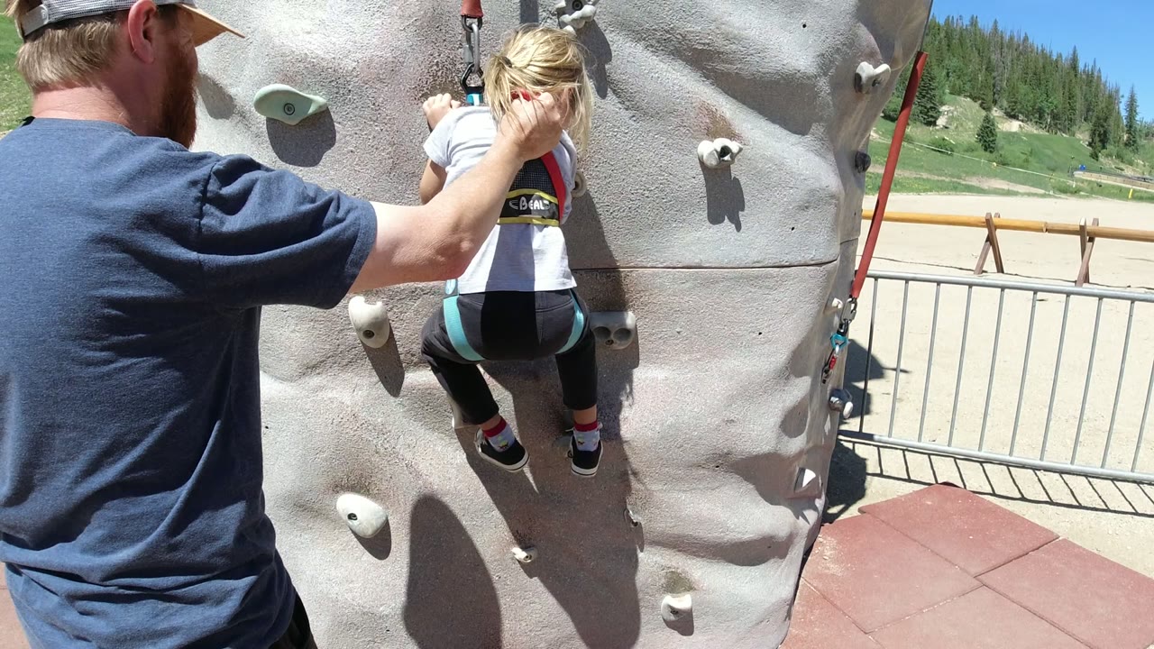 Adult helps a child climb an outdoor artificial rock wall.