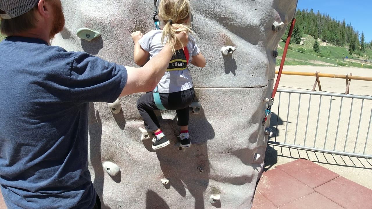 At 0:01 in Winter Park, Colorado, the child has moved higher on the gray climbing wall while the adult stays close with one hand near the child's back.