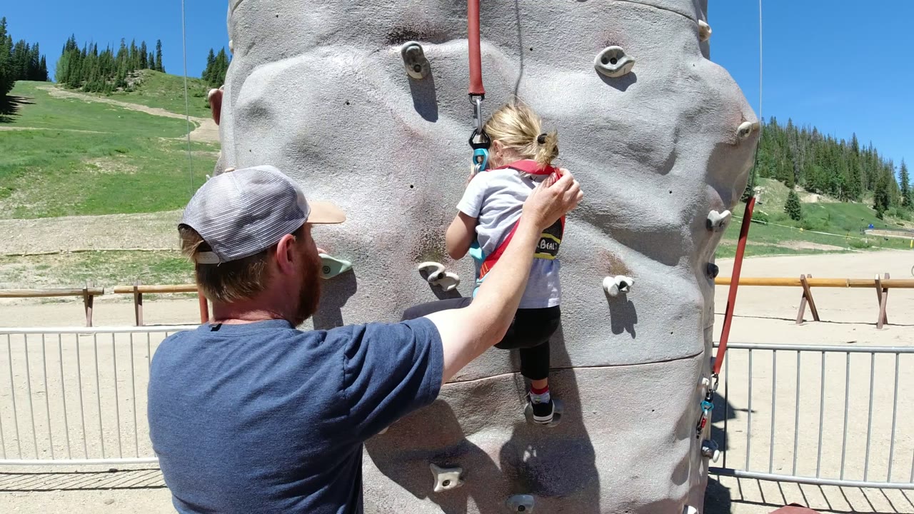 A man in a blue shirt reaches toward a harnessed little girl on an artificial climbing wall in Winter Park, Colorado, with green slopes and pine trees behind them.