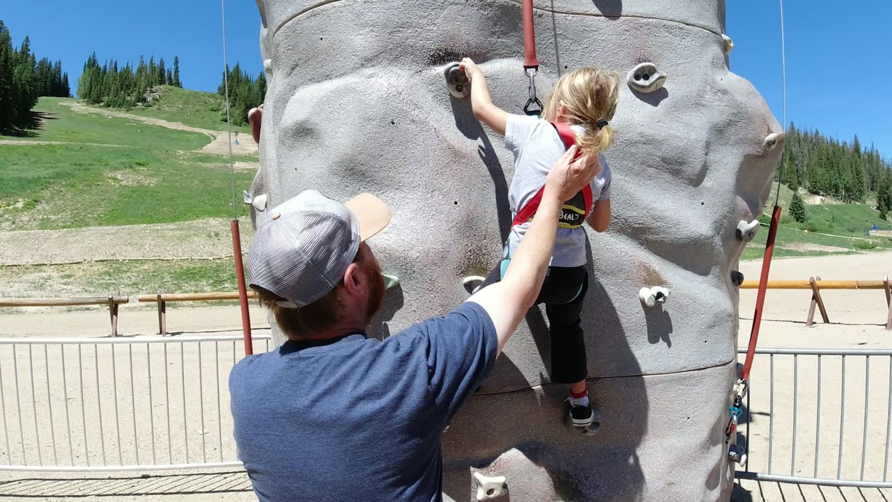 The little girl reaches to a higher hold on the gray climbing wall while the man keeps a hand near her back in the Winter Park recreation area.
