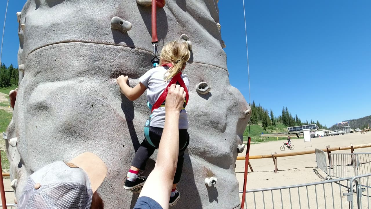 At Winter Park, Colorado, the girl shifts right on the artificial rock wall, with the adult's raised hand near her harness and a small bicycle visible in the background.