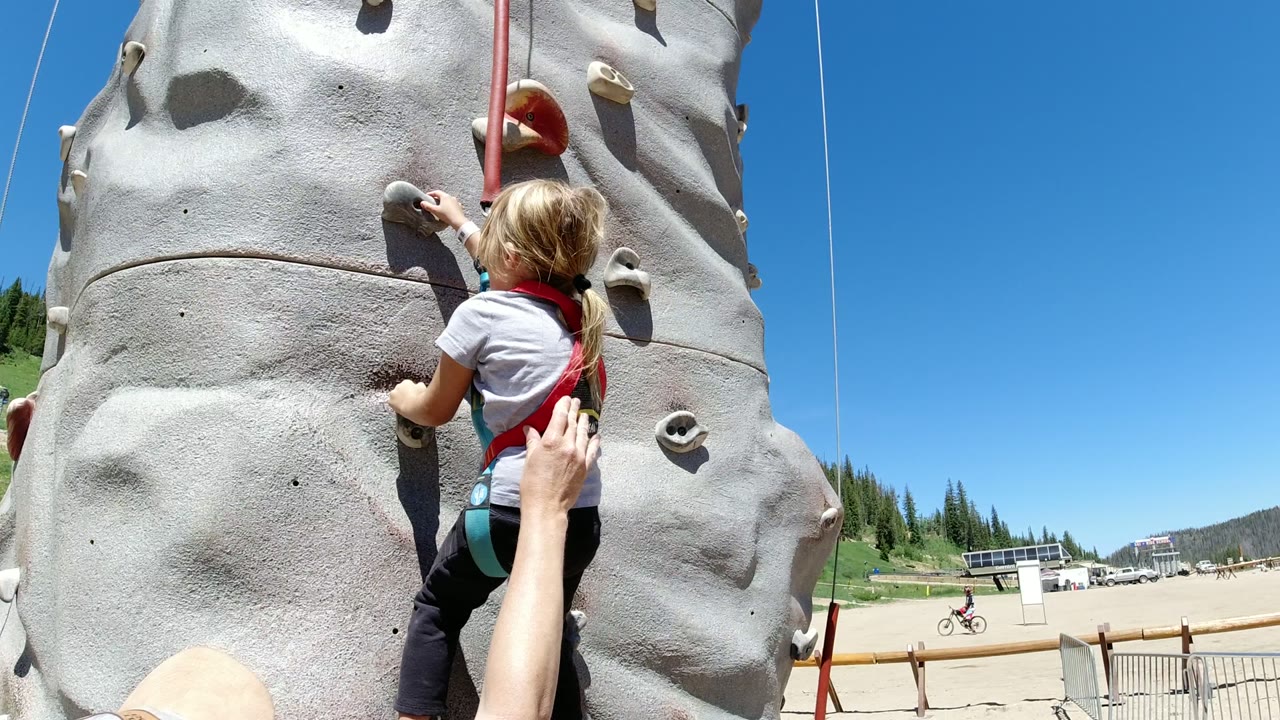 A young girl in a harness begins climbing a gray artificial rock wall in Winter Park, Colorado, with an adult hand near her back and blue sky above.