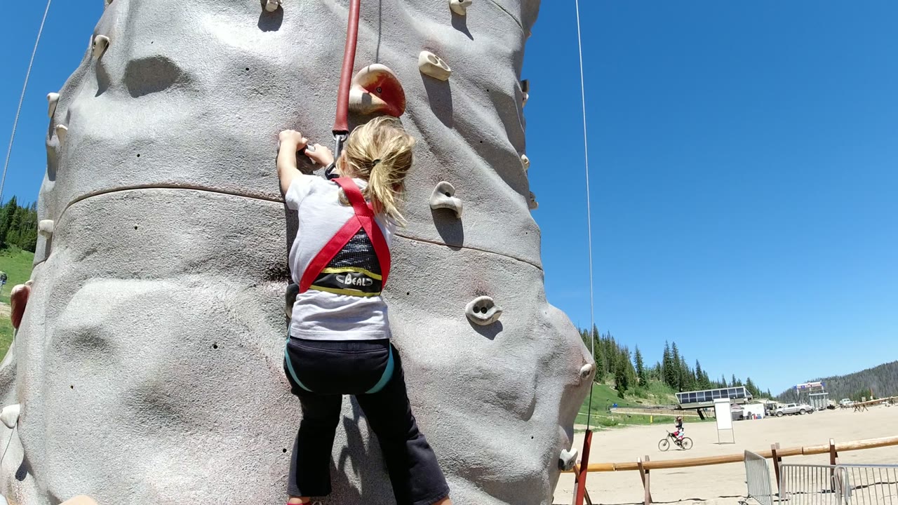 The girl reaches upward on the gray climbing wall in Winter Park, Colorado, with her harness straps visible and pine trees and open ground in the background.