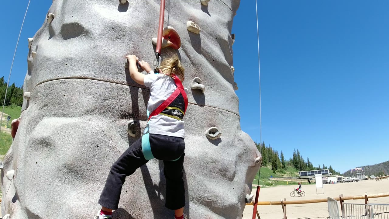 The child reaches higher on the artificial rock wall in Winter Park, Colorado, while a red belay rope hangs above and the mountain recreation area remains visible at frame right.