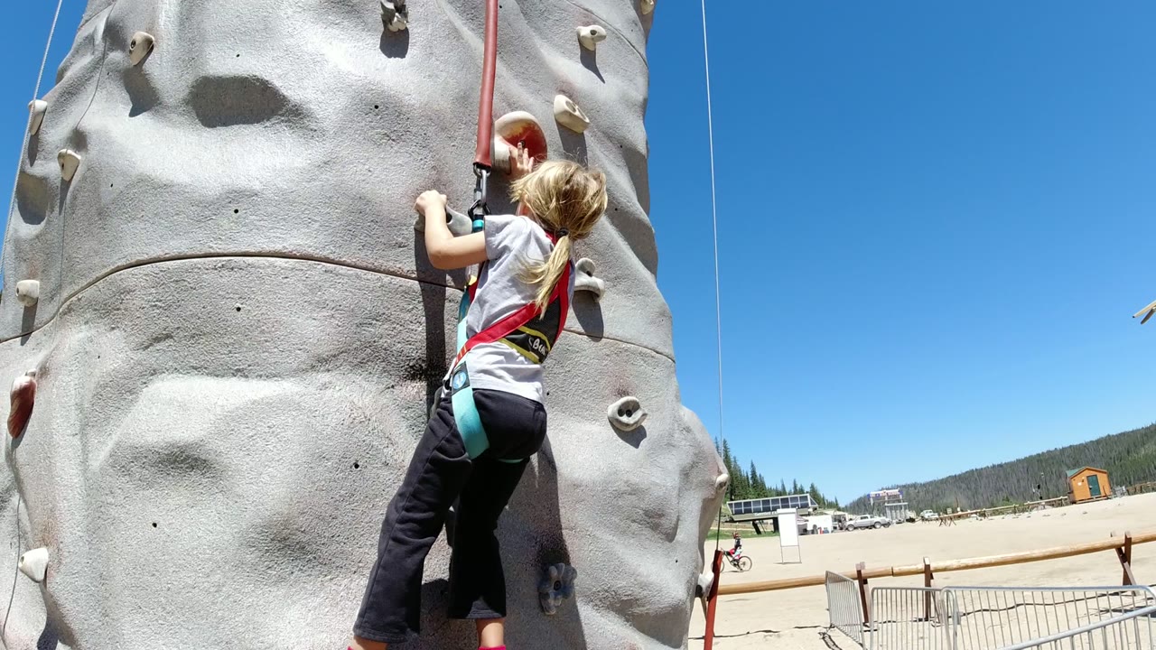 The child reaches toward a handhold on the gray climbing wall in Winter Park, Colorado, while a vertical rope and open recreation area remain visible to the side.