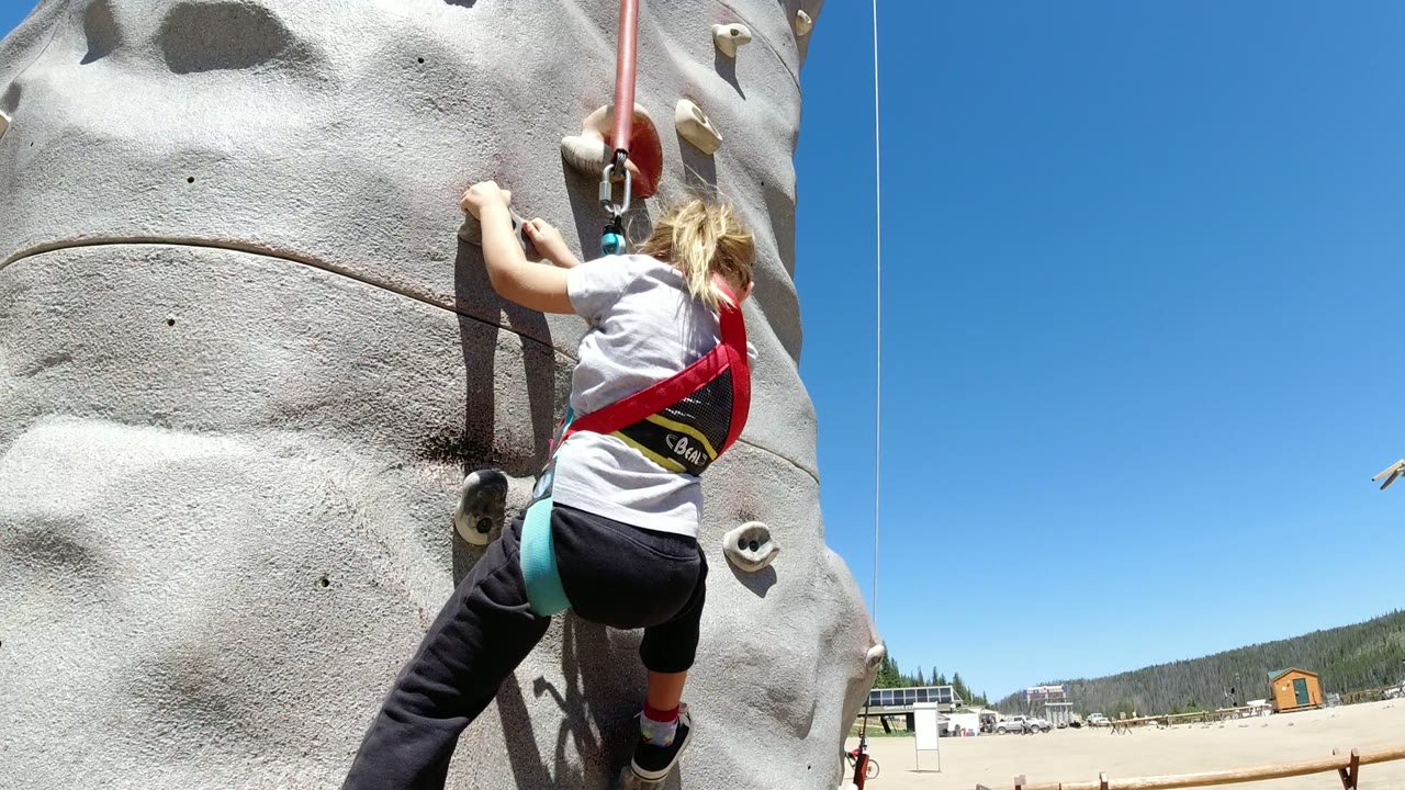 A young girl in a red harness reaches for holds on a gray artificial climbing wall in Winter Park, Colorado, with blue sky and open ground behind her.