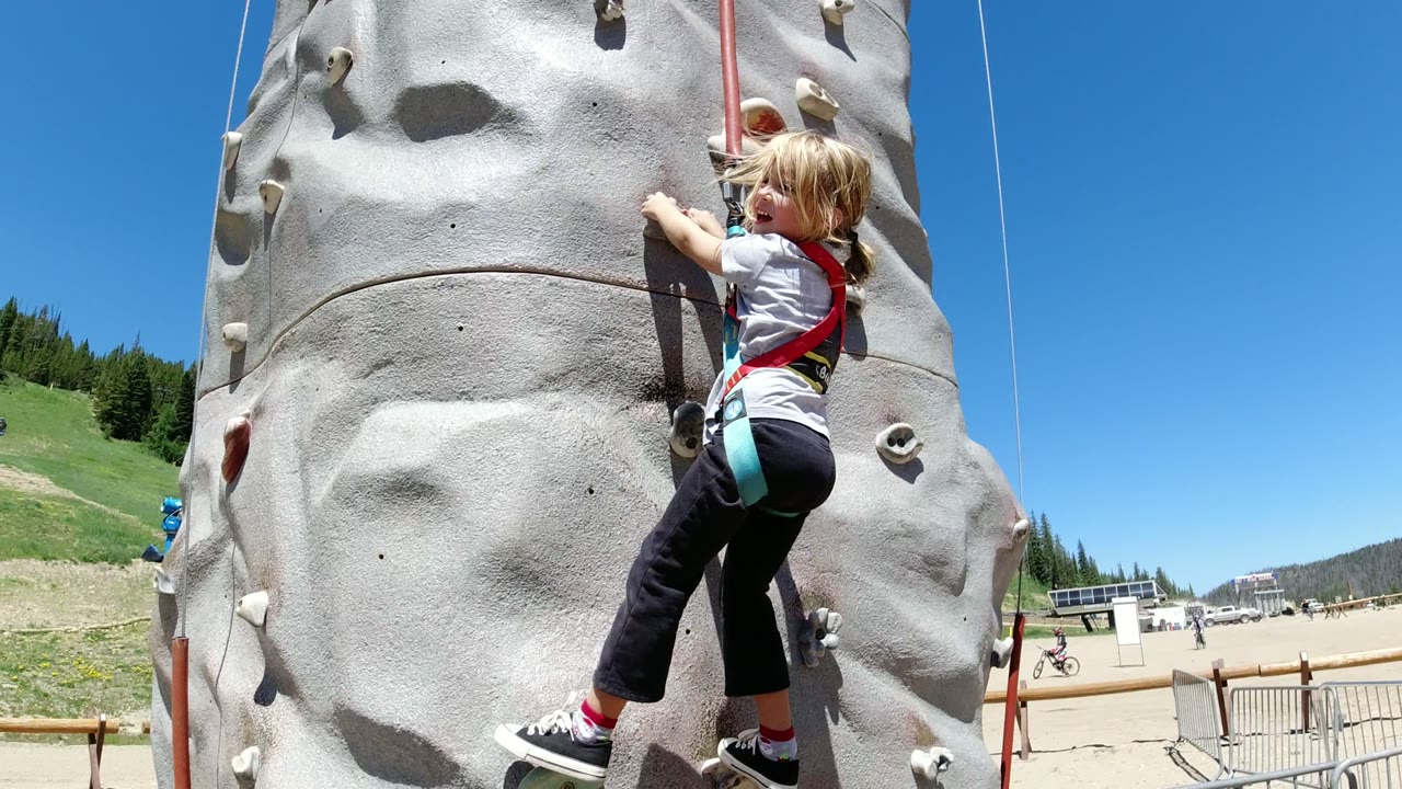 The child reaches across the gray climbing wall while suspended in a harness, with fencing, open paved ground, and distant slopes visible in Winter Park, Colorado.