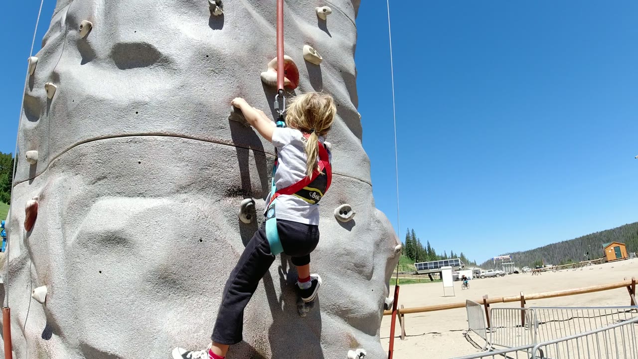 Young girl climbing a gray outdoor rock wall with a safety harness.
