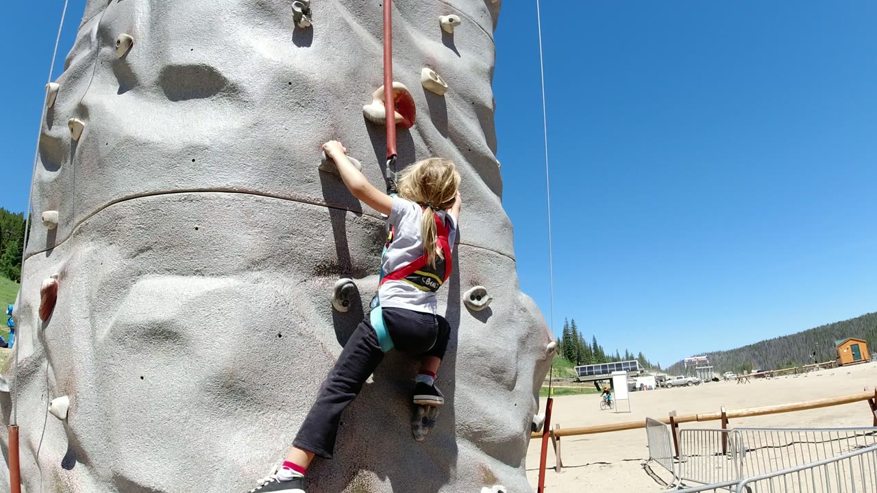 The girl moves higher on the Winter Park climbing wall, stretching her right arm toward a hold while the belay rope hangs beside her.