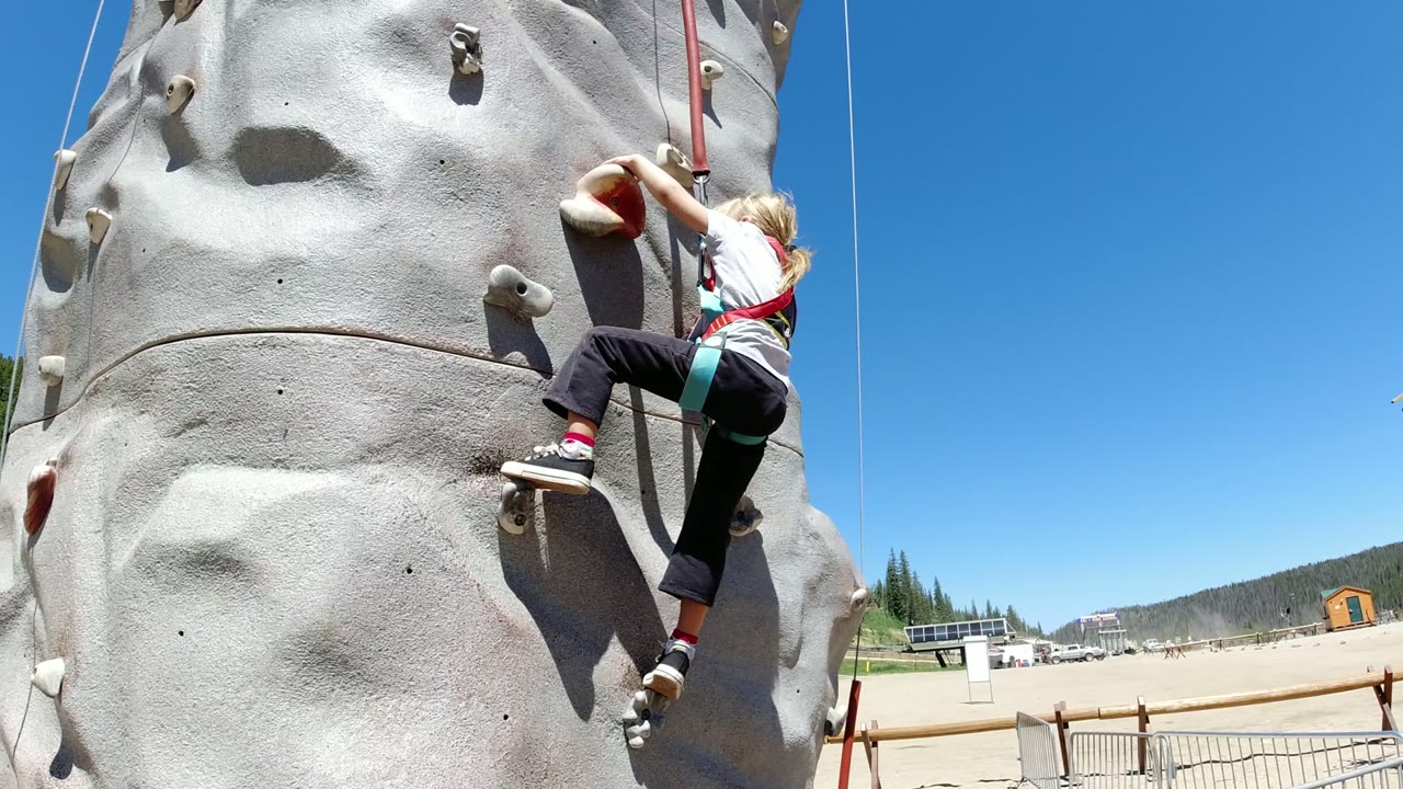 One second in, the girl extends toward a higher hold on the gray climbing wall, with the open blue sky and low mountain horizon visible in Winter Park, Colorado.