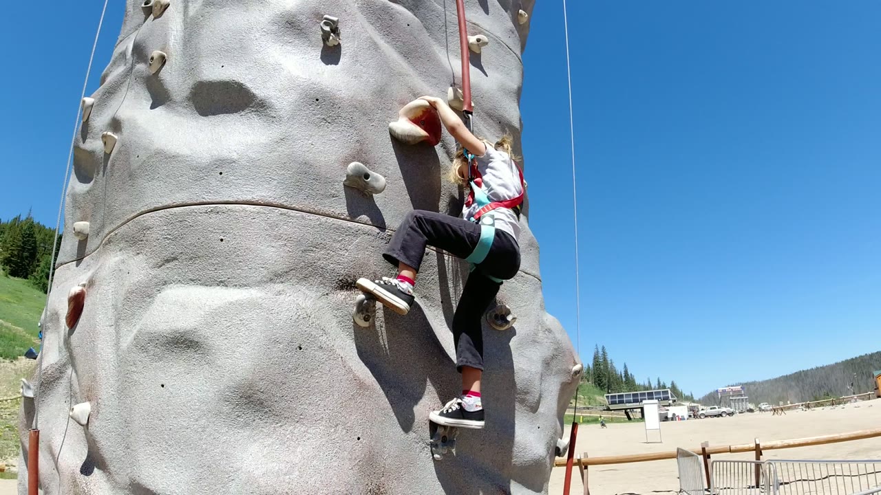 A young girl in a harness climbs low on a gray synthetic rock wall in Winter Park, Colorado, with a blue sky and mountain recreation area visible to the right.