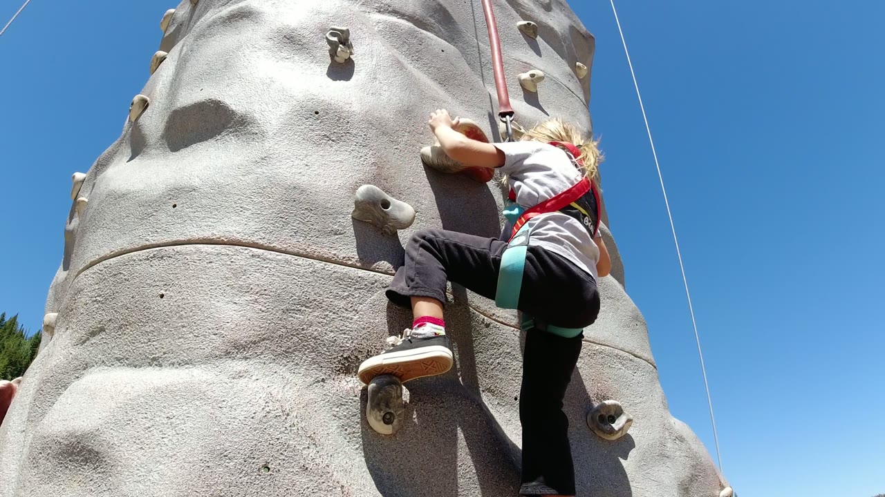 At the start of the Winter Park, Colorado clip, a harnessed child reaches upward on a gray artificial climbing wall with safety ropes against a clear blue sky.