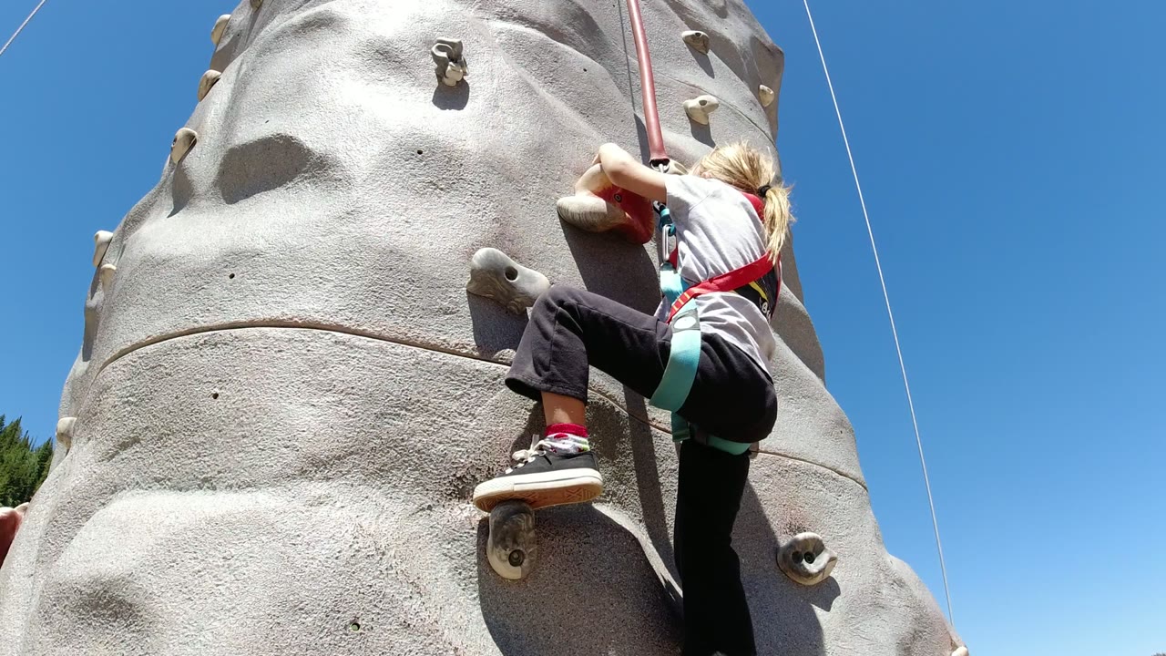 One second in, the child has one foot planted on a lower hold and one knee raised while climbing the gray wall in Winter Park, Colorado.