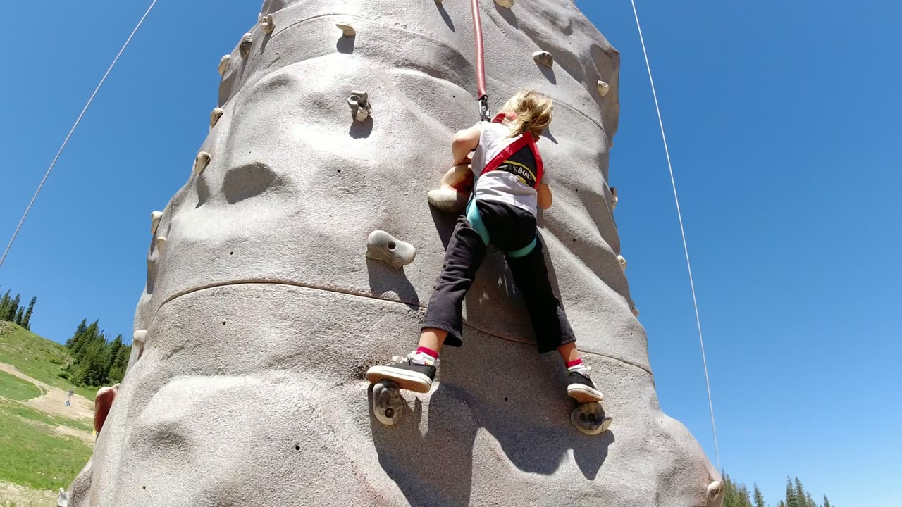 Girl climbing an outdoor rock wall with safety ropes against blue sky.