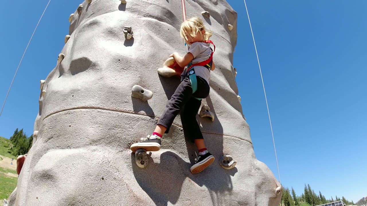 Young girl climbing an outdoor rock wall under a blue sky.