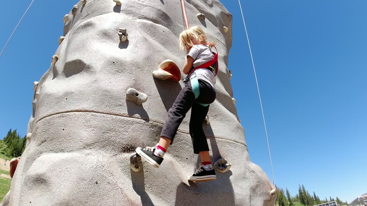 The young climber remains close to the gray wall in Winter Park, Colorado, stepping between holds while the red safety harness and rope are visible.